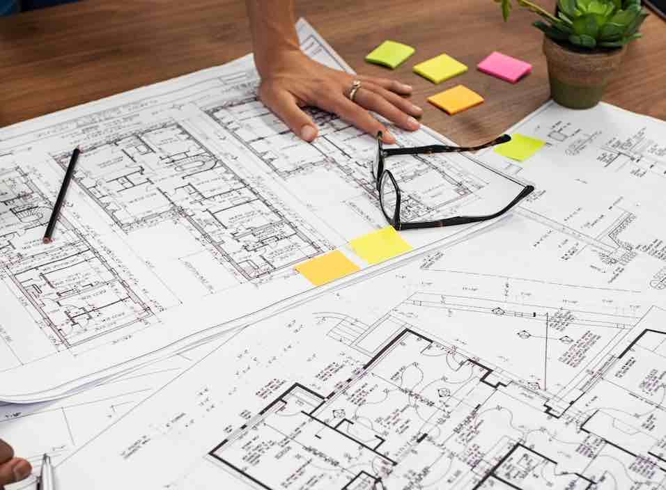 Closeup of multiethnic hands on blueprints at architects office. Team of designer and engineers working together on new residence complex. Top view of blue prints layout of house and buildings on wooden table.